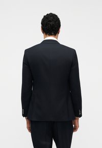 Άνδρας με κοντά σγουρά μαλλιά, φοράει σκούρο  σακάκι και παντελόνι, φαίνεται από πίσω με φόντο μια απλή φωτεινή επιφάνεια.