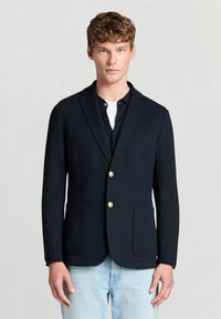 Navy blazer with a tailored fit, single-breasted design, gold buttons, and two front pockets, worn over a black shirt and light jeans.