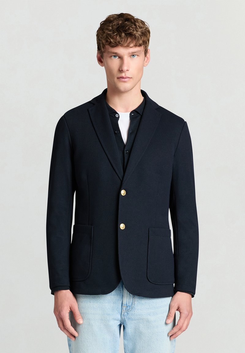 Navy blazer with a tailored fit, single-breasted design, gold buttons, and two front pockets, worn over a black shirt and light jeans.