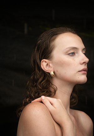 Gouden, textuurgeometrische oorringen gedragen door een persoon met nat, golvend haar, waarbij een schouder en hand zichtbaar zijn. Zacht licht en een neutrale achtergrond.