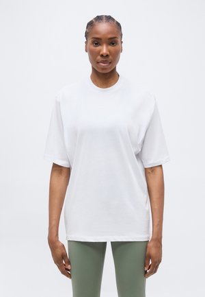 Γυναίκα με πλεξούδες, που φοράει ένα απλό λευκό φαρδύ μπλουζάκι και ανοιχτοπράσινο κολάν, στέκεται μπροστά σε λευκό φόντο.