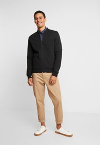 Sort zip-up jakke med ribbet tekstur og opretstående krave, båret over en marineblå skjorte. Kombineret med beige chinos, der er rullet op, og hvide sneakers.