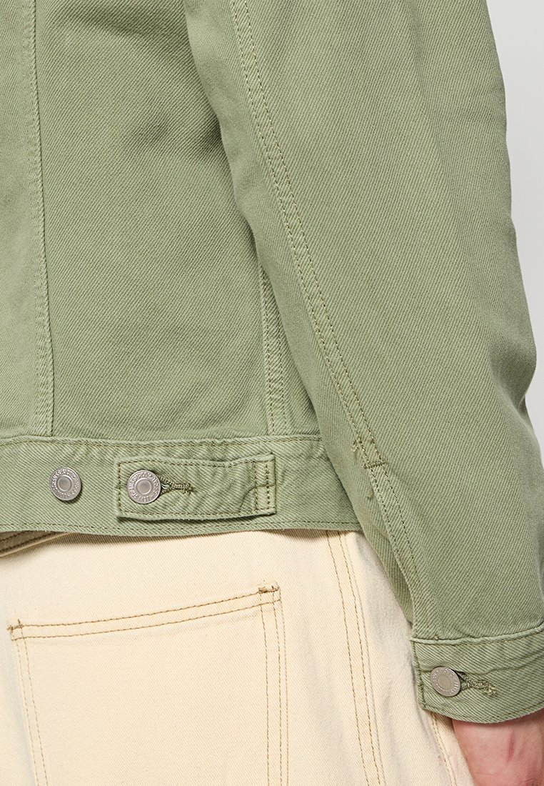 Giubbotto di jeans verde chiaro con chiusure a pressione metalliche, cuciture a vista e vestibilità strutturata. Tessuto morbido e testurizzato con dettagli laterali.