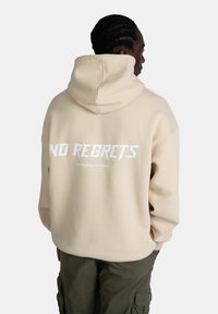 Felpa oversized beige con cappuccio regolabile. Presenta una grande scritta bianca "NO REGRETS" sul retro e un testo più piccolo sotto. Tessuto morbido, vestibilità casual.