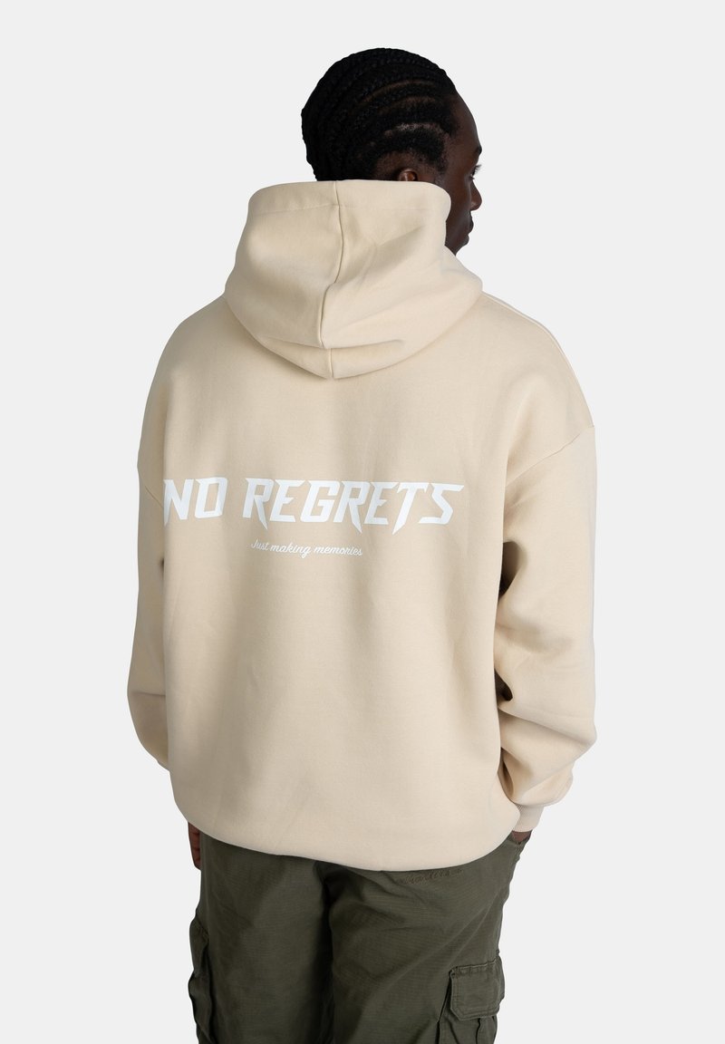 Felpa oversized beige con cappuccio regolabile. Presenta una grande scritta bianca "NO REGRETS" sul retro e un testo più piccolo sotto. Tessuto morbido, vestibilità casual.