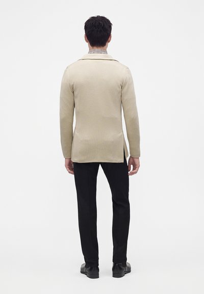 Homme portant une veste beige, une chemise à carreaux, un pantalon noir et des chaussures noires, debout, de dos sur un fond blanc uni.