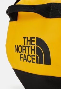 Yellow waterproof bag with a black bottom, featuring a large black logo, structured shape, and black webbed handle.