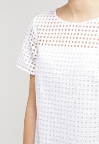 White short-sleeve top with a square-patterned mesh fabric showing skin underneath on upper chest and sleeves.