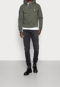 Felpa verde oliva con tasca frontale e cappuccio con lacci, indossata sopra una camicia bianca; abbinata a jeans neri slim e stivali chelsea neri.