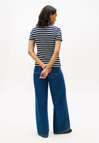 Nainen seisoo selin kameraan, pukeutuneena laivastonsiniseen ja valkoiseen raidalliseen paitaan ja leveälahkeisiin sinisiin farkkuihin, kädet ristissä selän takana.