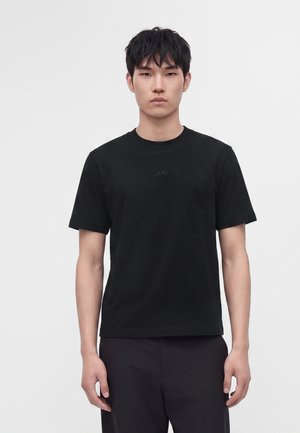Ung mand med kort sort hår iført en ensfarvet sort t-shirt med korte ærmer og sorte bukser, stående mod en lys grå baggrund.