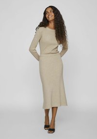 Robe pull à manches longues en beige avec une texture côtelée, ajustée à la taille, s'évasant vers l'ourlet, associée à des sandales à talons noires.