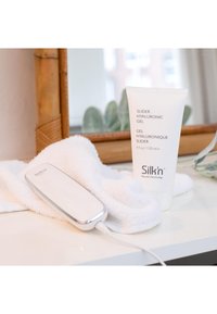 Hair removal device in white with silver accents, placed on a white towel beside a tube of hyaluronic gel. Minimalist and sleek design.