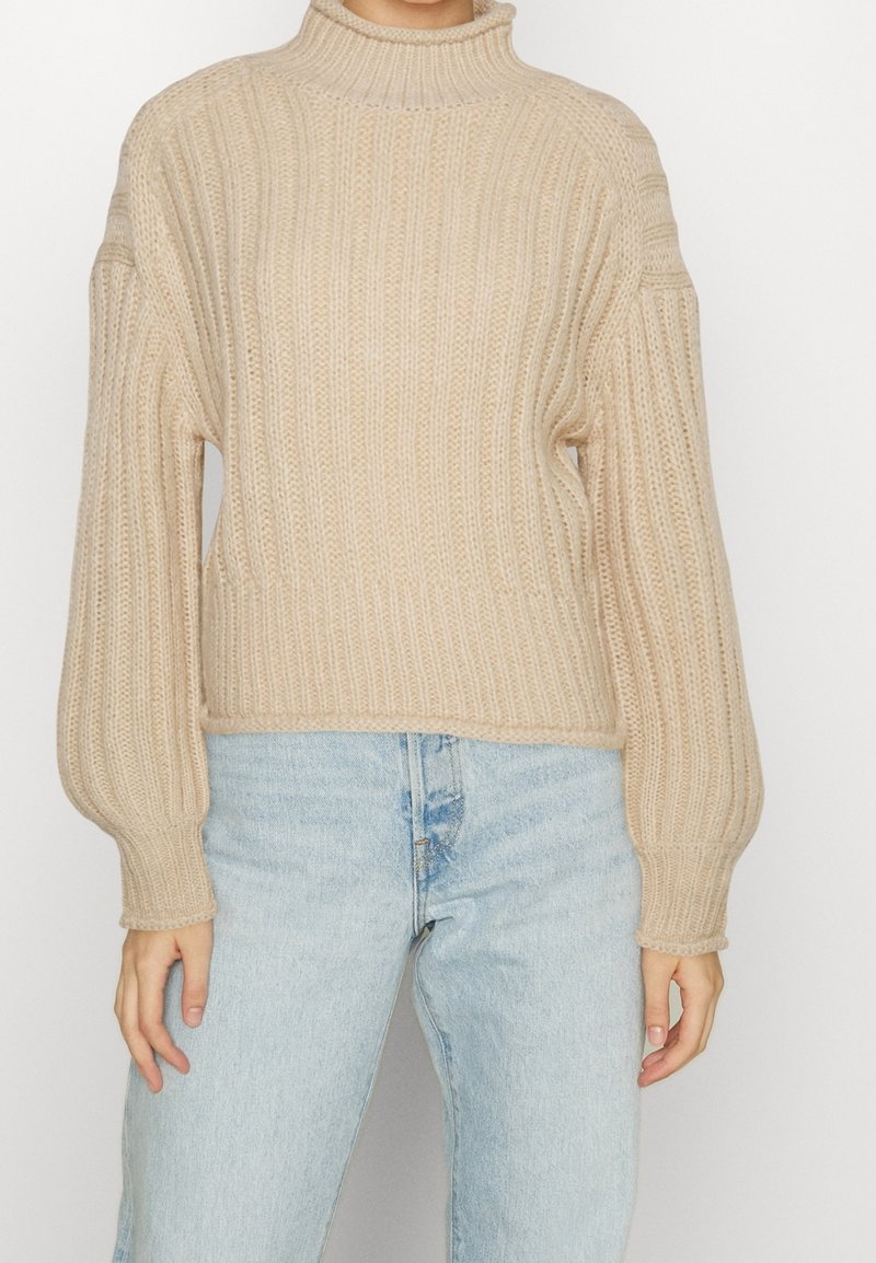 Personne portant un pull à col roulé en grosse maille beige avec des manches bouffantes et un jean taille haute bleu clair sur un fond uni.