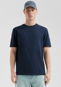 Granatowa koszulka z krótkim rękawem z ukośnym przeszyciem; wykonana z bawełny. Połączona z jasnoniebieskimi dżinsami i niebieską czapką.