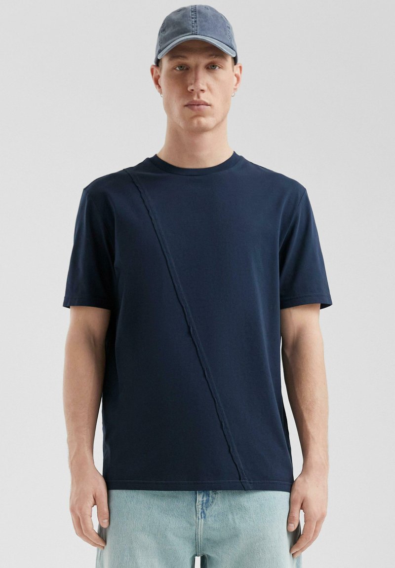 Granatowa koszulka z krótkim rękawem z ukośnym przeszyciem; wykonana z bawełny. Połączona z jasnoniebieskimi dżinsami i niebieską czapką.