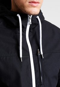 Black hooded jacket with white drawstrings and zipper, featuring a front pocket with a zip closure and a smooth, matte texture.