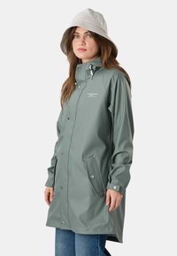 Žena v dlhom dažďovom kabáte šalviovo zelenej farby so zapínaním na patentky a v bielom klobúku typu bucket, stojaca pred jednoduchým pozadím.