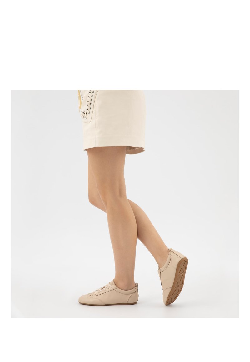 Frau trägt beige flache Schnürschuhe und passenden kurzen Rock, steht mit einem leicht angehobenen Fuß.