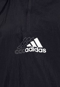 Zwarte jas met een kwartritssluiting, gemaakt van lichtgewicht materiaal, met een opvallend wit Adidas-logo linksonder.