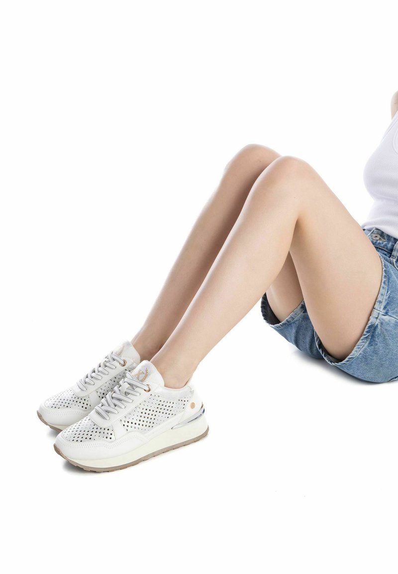 Zapatillas perforadas blancas con suela acolchada y cordones gris claro, que presentan detalles dorados y un diseño de punta redondeada.