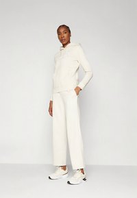 Krémová mikina s kapucňou a predným vreckom, v kombinácii so širokými nohavicami. Obe sú vyrobené z mäkkej látky a majú jemné logo.