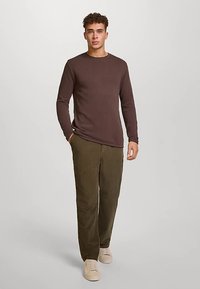 Braunes Langarmhemd, olivgrüne Hose, beige Sneakers. Das Hemd hat einen Rundhalsausschnitt; die Hose ist gerade geschnitten mit Seitentaschen.