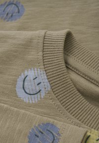Beige stoffen shirt met een geribde halslijn, bedrukte glimlachende gezichten in blauw en groen, en een zachte, gestructureerde textuur.