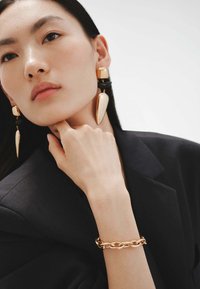 Pendientes de oro con formas alargadas, un detalle negro y una pulsera de cadena de oro. El sujeto lleva un blazer oscuro, presentando un fondo neutro.