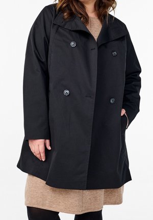 Frau trägt einen schwarzen, doppelreihigen Mantel über einem beigen Kleid, mit einer Hand in der Tasche und der anderen locker an der Seite.