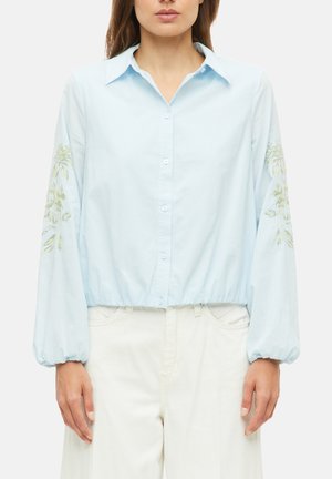 Frau trägt eine hellblaue Bluse mit Puffärmeln und Blumenstickerei, kombiniert mit hoch taillierten weißen Hosen.