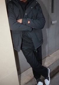Veste rembourrée noire avec capuche, dotée de poches zippées et d'un logo rouge et jaune sur la manche. Portée par-dessus un sweat-shirt noir et un pantalon.
