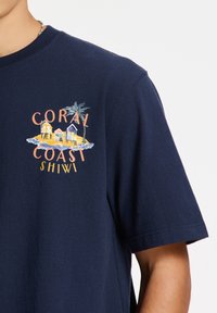 Camiseta de algodón azul marino con un estampado gráfico de coloridas casas de playa y palmeras, con el texto "CORAL COAST SHIWI" debajo.