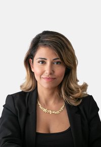 Portrait einer Frau mit schulterlangen, welligen Haaren. Sie trägt ein schwarzes Blazer und eine goldene geometrische Halskette. Der Hintergrund ist schlicht weiß.