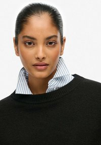 Black knitted sweater with a high neck, layered over a light blue and white striped collared shirt. Smooth texture and casual design.