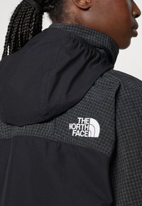 Fekete dzseki rácsos mintázatú résszel a vállán. Az „THE NORTH FACE” logó látható a mellkason. Könnyű, szövet textúra.