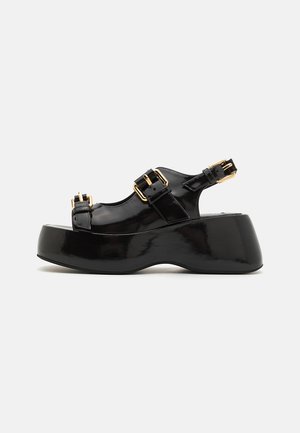 Zwarte patenten leren plateau sandalen hebben een chunky hak, twee verstelbare gespstraps en een gladde, glanzende afwerking.