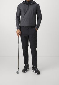 Pánský černý lehký pulovr s kapucí a čtvrtinovým zipem, v kombinaci s černými kalhotami a černými sportovními botami, který drží golfovou hůl.