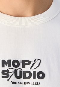 Camiseta blanca de manga corta con cuello redondo de canalé y texto negro en negrita: "MO’PD STUDIO" y "Estás INVITADO." Textura de tela suave.