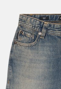 Blugi din denim albastru cu croială lejeră, având cusături de culoare cupru, închidere cu nasturi și două buzunare frontale. Eticheta spune "BAGGY ALEX."
