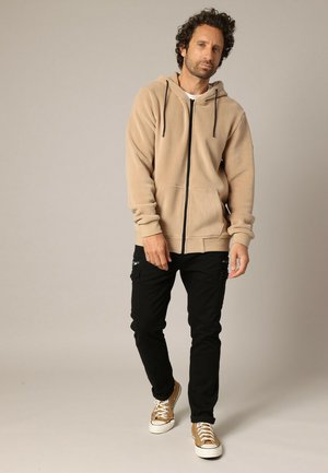 Haut à capuche zippé beige avec un cordon de serrage, deux poches avant, associé à un pantalon cargo noir et des baskets beige. Texture de tissu lisse.