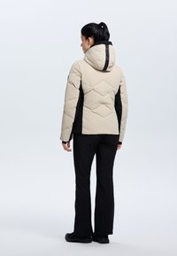 Beige gepolsterte Jacke mit schwarzen Seiten und Kapuze, mit einem gesteppte Design und einem vertikalen schwarzen Streifen auf dem Rücken. Schwarze Hose und Schuhe.