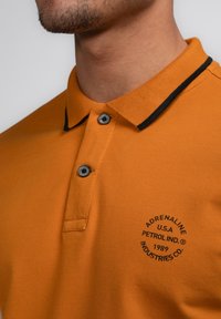 Oranžové polo tričko z texturované tkaniny, s límcem s černým lemem, dvěma šedými knoflíky a kruhovým černým logem na hrudi.