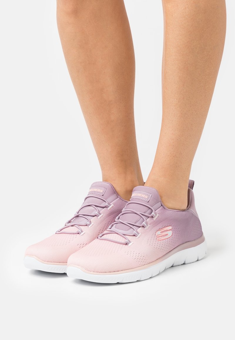 Jambes portant des baskets Skechers avec un dégradé rose et violet et des semelles blanches, debout sur un sol gris clair.