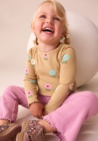 Brun revet langærmet skjorte med farverige blomsterprint, kombineret med lyserøde fløjlsbukser med vidde. Fodtøjet har blomsterdetaljer.