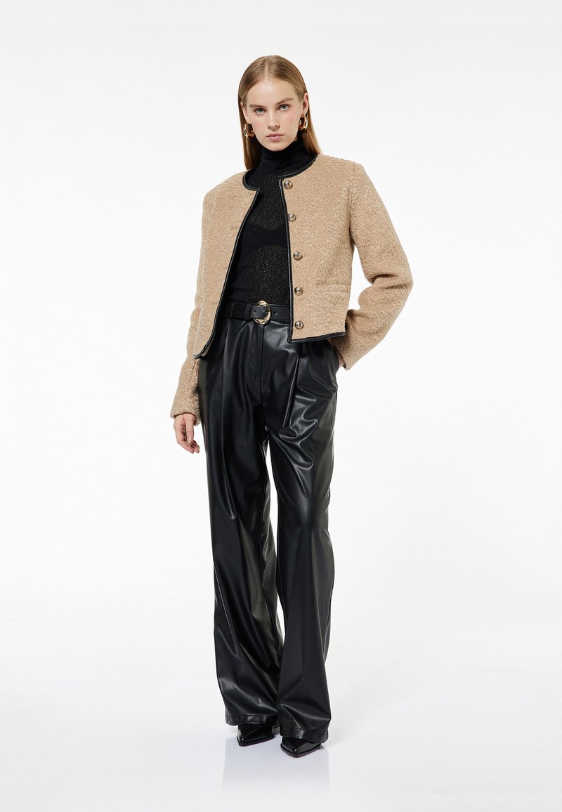 Beżowa kurtka o fakturze z okrągłym dekoltem, wyeksponowana na otwarto, z czarnymi zdobieniami i dużymi guzikami. Czarne skórzane spodnie z wysokim stanem dopełniają całość.