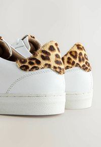 Baskets blanches avec semelles en caoutchouc texturées et accents de talon en fausse fourrure à imprimé léopard sur un fond clair.