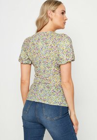 Blusa floreale a maniche corte in morbido tessuto; base gialla con motivo floreale in rosa, blu e verde. Design aderente con dettagli arricciati sulle spalle.