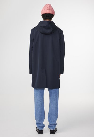 Marinblå vattentät kappa med huva, enkel ventil bak, matchad med ljusblå jeans och en rosa stickad mössa. Slät textur, minimalistisk design.