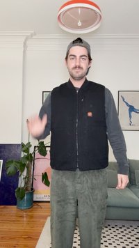 Colete preto com fecho zip sobre uma camisa de manga comprida escura, combinado com calças de veludo cotelê verdes. Uma luz redonda no teto está visível acima.